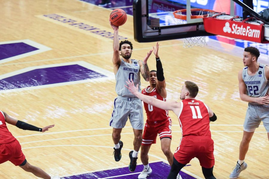 Boo Buie, wearing a grey, number zero jersey, jumps with the ball in his hand to score a basket, surrounded by defenders in red, Wisconsin jerseys.