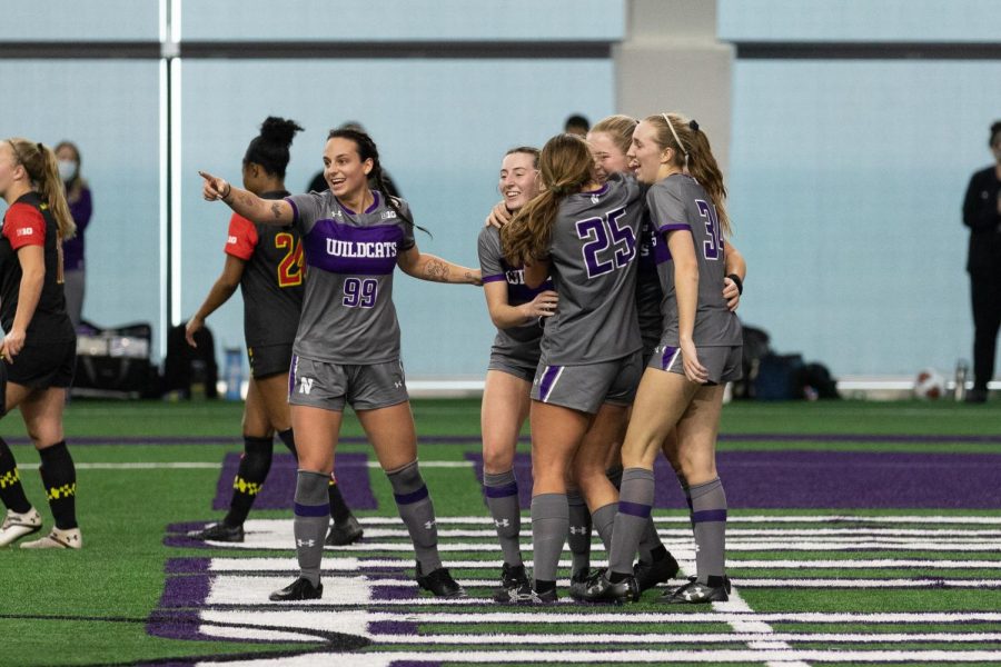 Women’s soccer players wearing gray and purple uniforms hug each other.