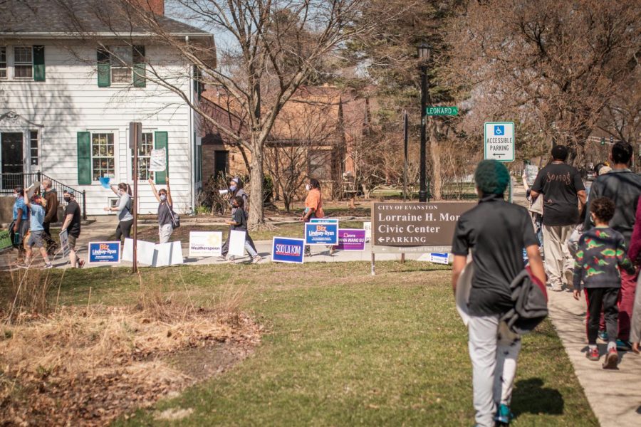 Participants march on the sidewalk past the sign for the Lorraine H. Morton Civic Center. Next to the Civic Center sign, there are several lawn signs for municipal election candidates, including ones promoting Elisabeth “Biz” Lindsay-Ryan, Soo La Kim, and Marquise Weatherspoon.