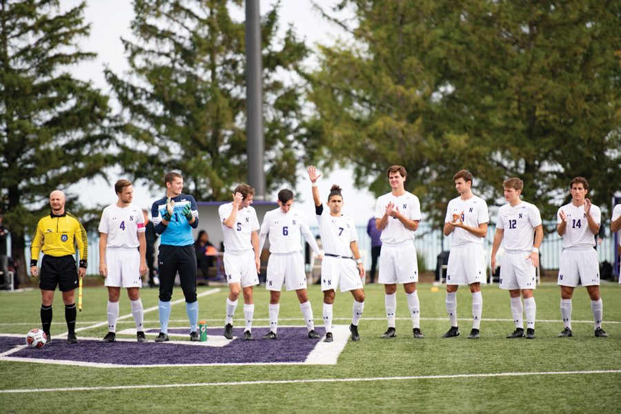 Players in white jersey stand in a line on green grass field with one in front of the others in the horizontal line, waving