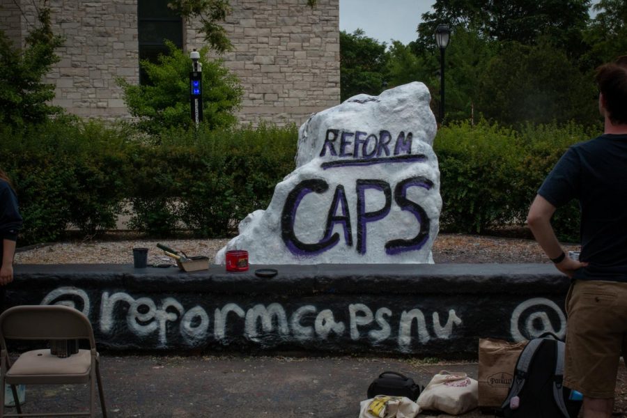 The Rock, painted in white, states “Reform CAPS.” A person in tan shorts and a navy shirt faces it. In the background are green shrubs, blue sky, and tan buildings.