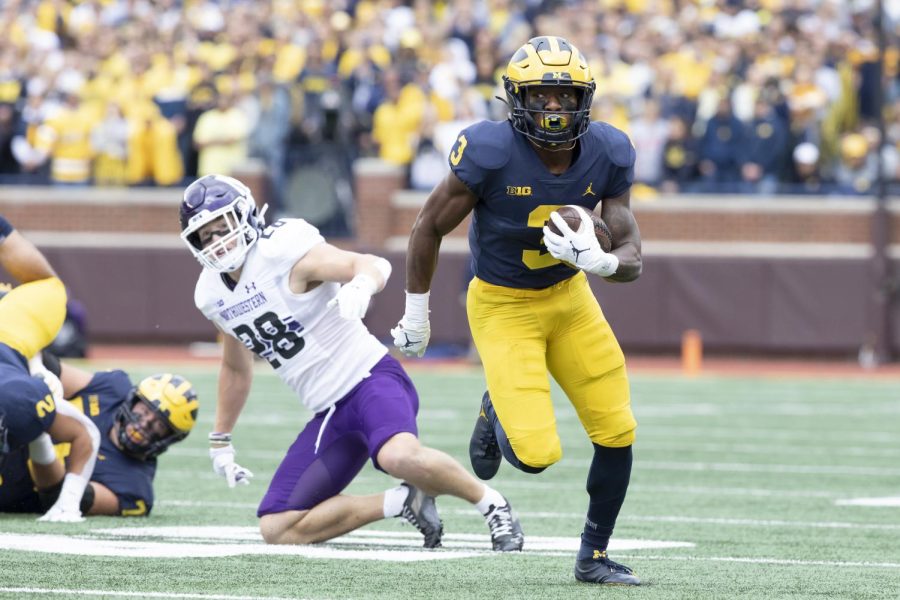 A Michigan player in a blue jersey runs with the ball along the white sideline. An NU player in a white jersey watches from the background, having fallen to the ground.