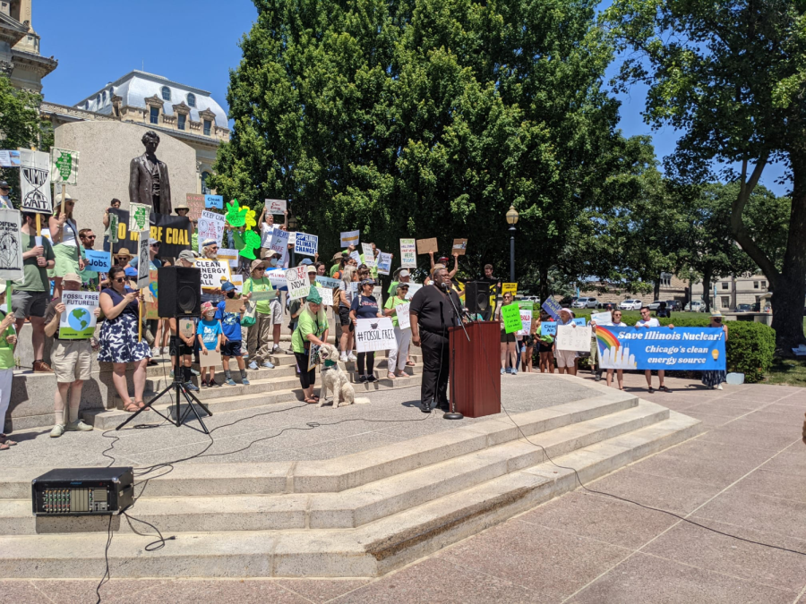 On steps and in front of a statue, a large crowd mostly dressed in green-and-blue holds pro-nuclear signs. A speaker in all black stands at the podium.