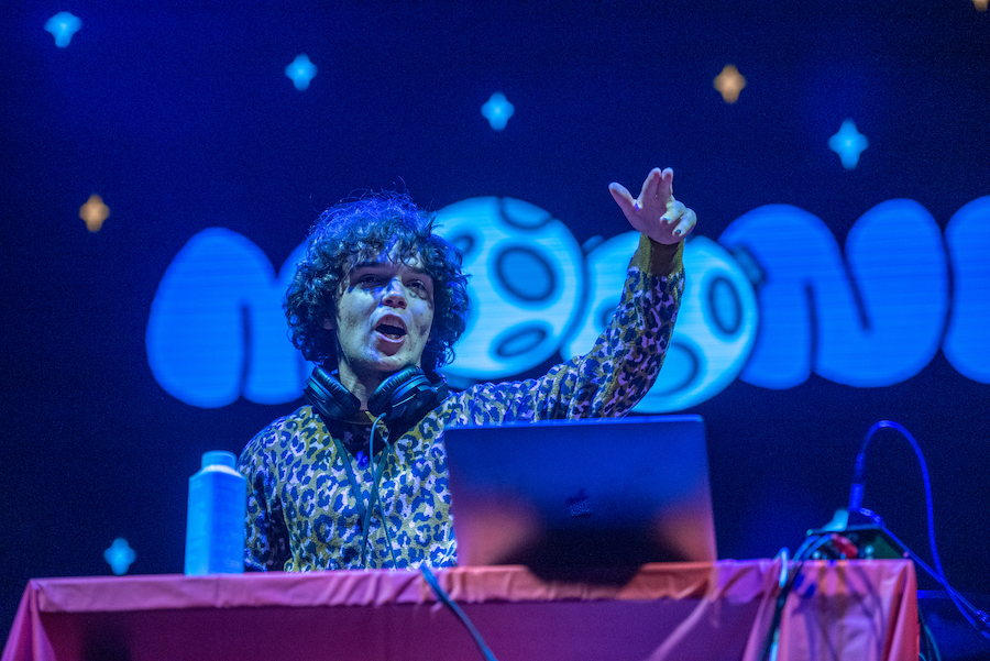 Tyler stands under blue light with his arm in the air, in front of an open laptop and headphones around his neck. He’s on stage in front of a screen with the word “Moondog” on it.