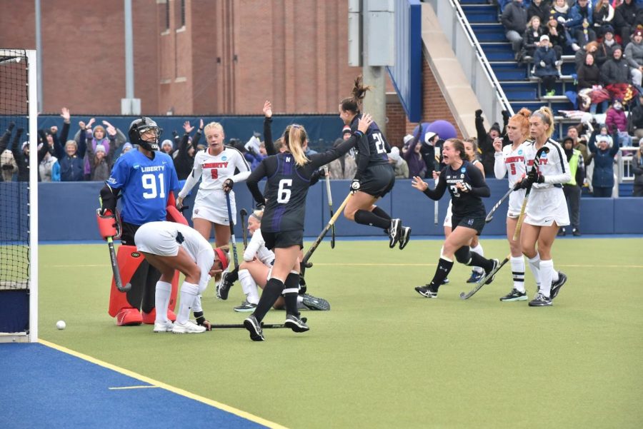 Three field hockey players dressed in black jump in celebration.