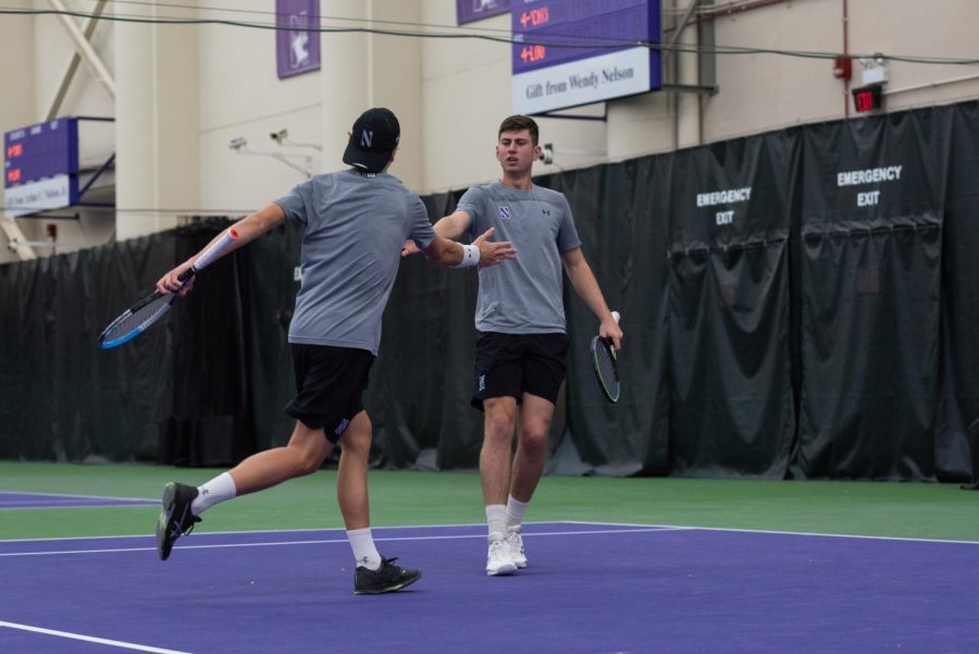 Two male tennis players, one with hat and black shoes and one with white shoes, high-five on purple and green court.