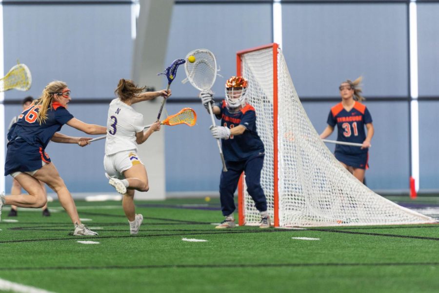 A woman in a white jersey shoots a goal toward a lacrosse goalie in blue jersey.