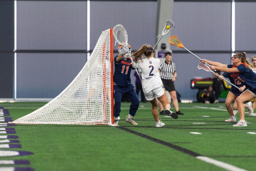 A woman playing lacrosse in a white jersey shoots a ball behind her back toward a lacrosse goal.