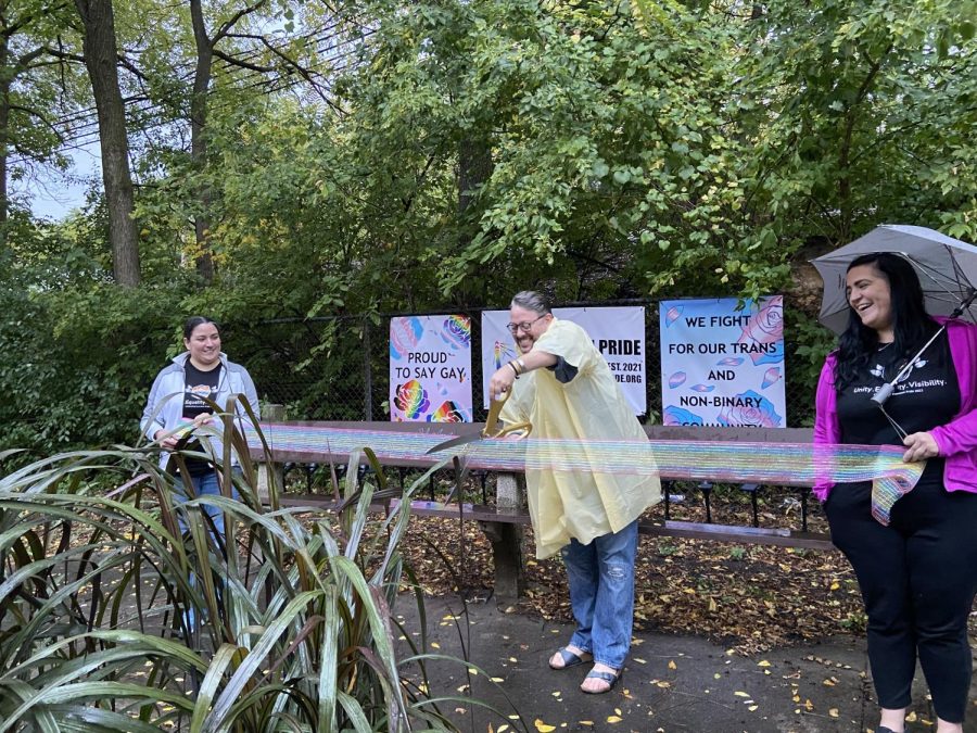 A man holding giant scissors snips a large, sparkly rainbow ribbon held by others on either side. Posters depicting pride flags and slogans hang along a fence in the background.