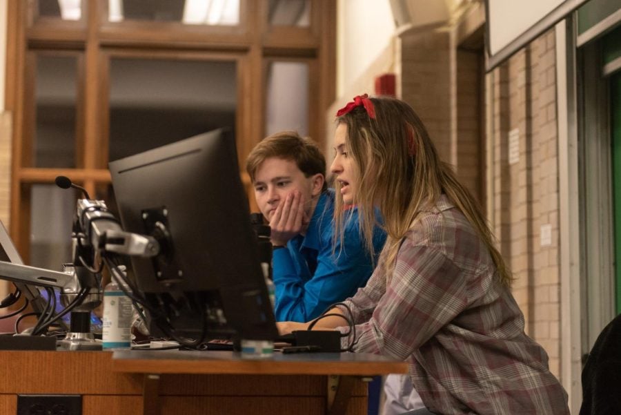 Two individuals, one in a blue shirt and another in a flannel, looking at a computer screen on top of a desk.