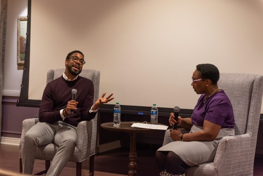 A man in a maroon sweater sits opposite a woman in a purple top. Holding a microphone in one hand, he reaches out another hand as he speaks.
