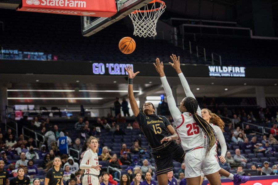 Basketball players, one in a black and gold jersey, and two in white and red jerseys, reach for the ball.