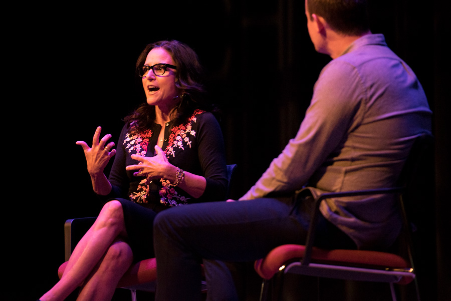 Julia Louis-Dreyfus sitting on a dark stage addressing an audience, sitting next to a man in a gray shirt.