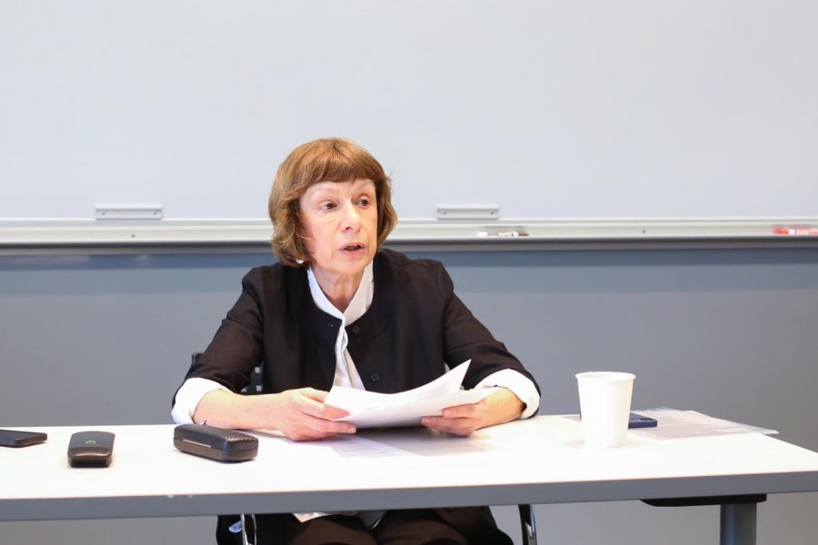 A woman sitting at a desk holds papers and speaks.