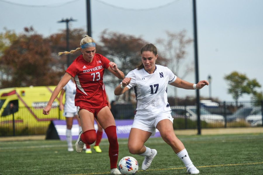 A person in a white uniform kicks a soccer ball away from another player in red.