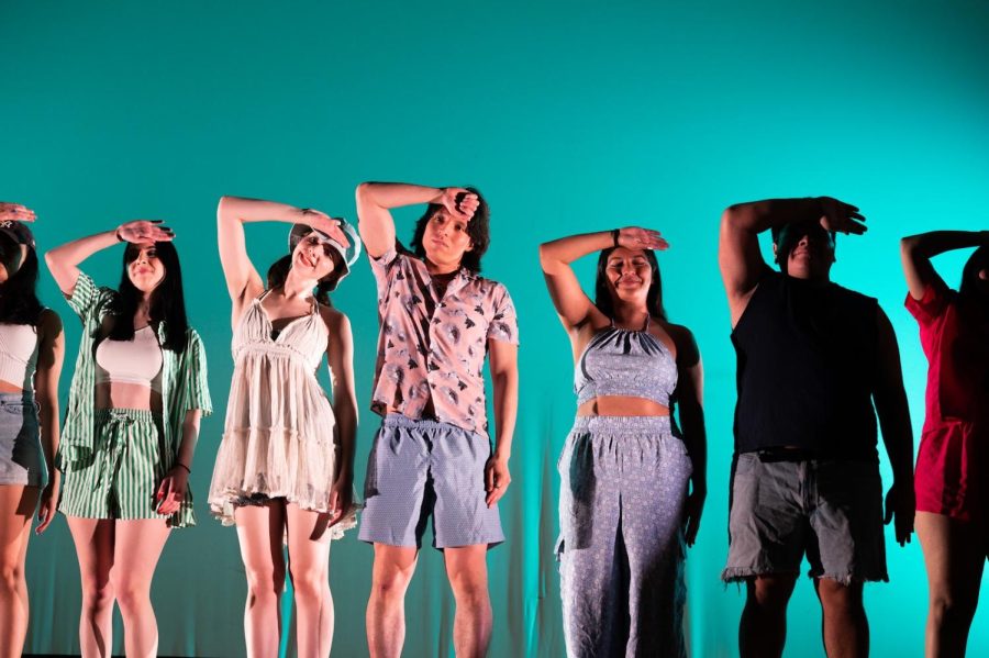 Dancers in beach attire stand against a blue backdrop with their arms across their foreheads.