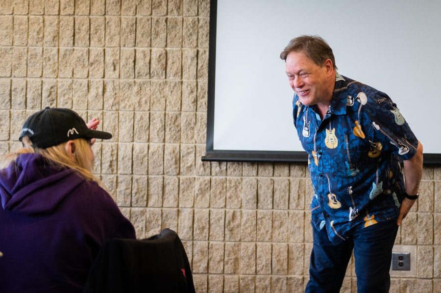 A student in a purple hoodie and black cap raises their left hand as a professor looks at them with a smile.