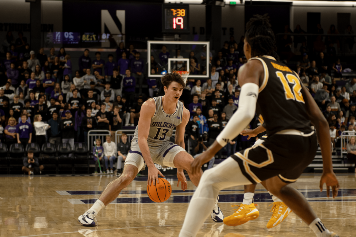 An athlete in a gray jersey dribbles a basketball between their legs.