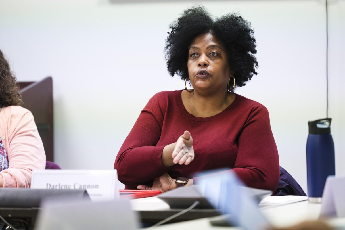Woman wearing a red long-sleeve shirt sits at a table behind a placard reading, “Darlene Cannon.”
