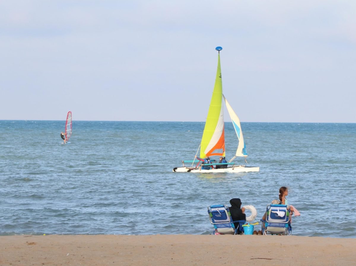 People sitting on beach chairs at the beach facing a sailboat on the water.