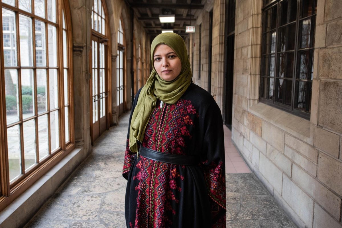 A woman wearing a black and red dress and a hijab stands in the hallway of a stone building.