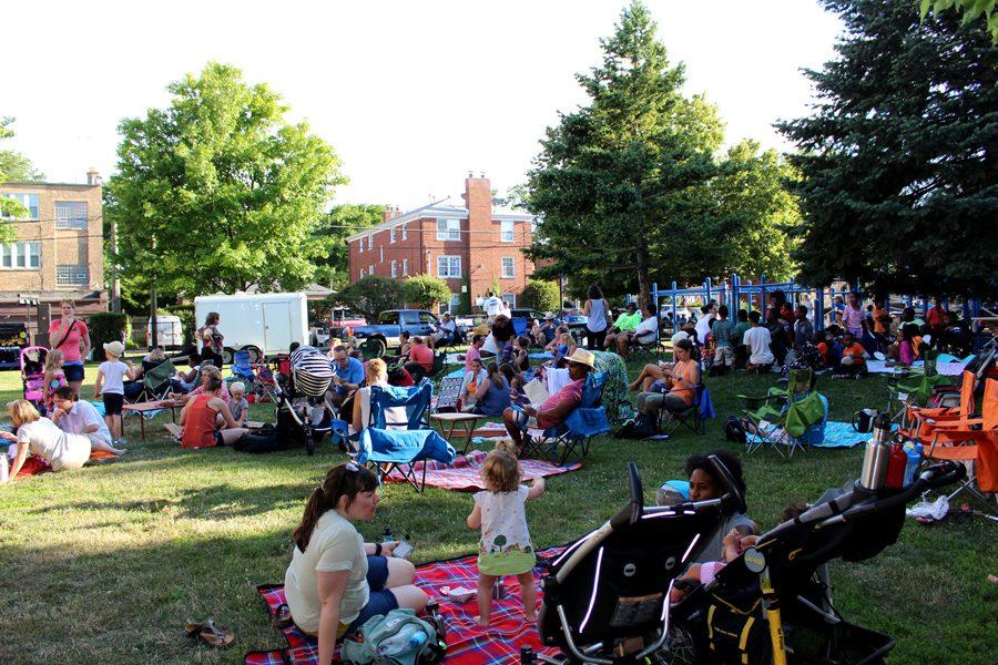 A crowd of people of all ages sit in an open park on blankets.